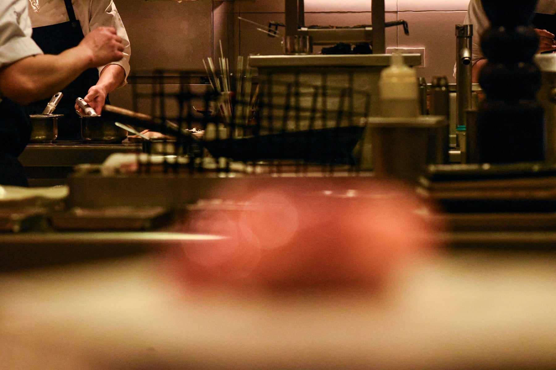 Chef en cuisine professionnelle, mains actives derrière un plan de travail, ambiance de service en cours.