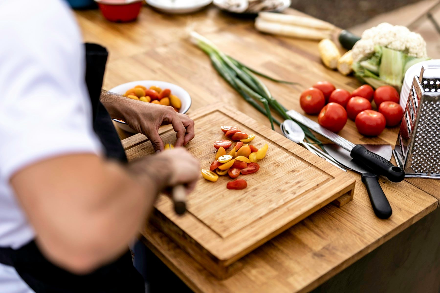 Chef en veste blanche découpant des tomates cerises sur une planche en bois, entouré de légumes frais et d'ustensiles.
